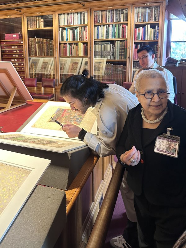 Someone looks closely at a book of prints on a book rest in a library. Meanwhile a woman turns a little away from the book herself