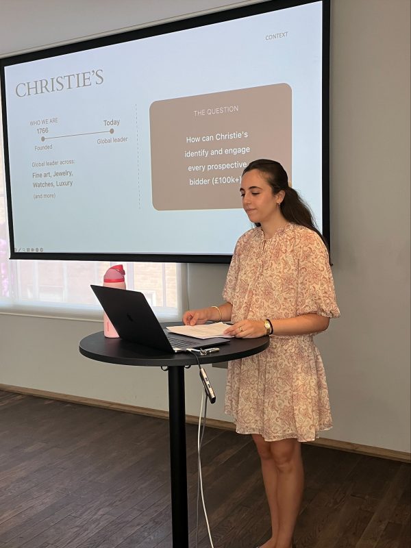 A student stands in front of a laptop at a podium. Behind her, a projector shows a slide on how Christie's can engage prospective bidders