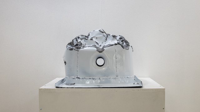 white metal object similar to a sink sits on a plinth displaying signs of violent destruction.