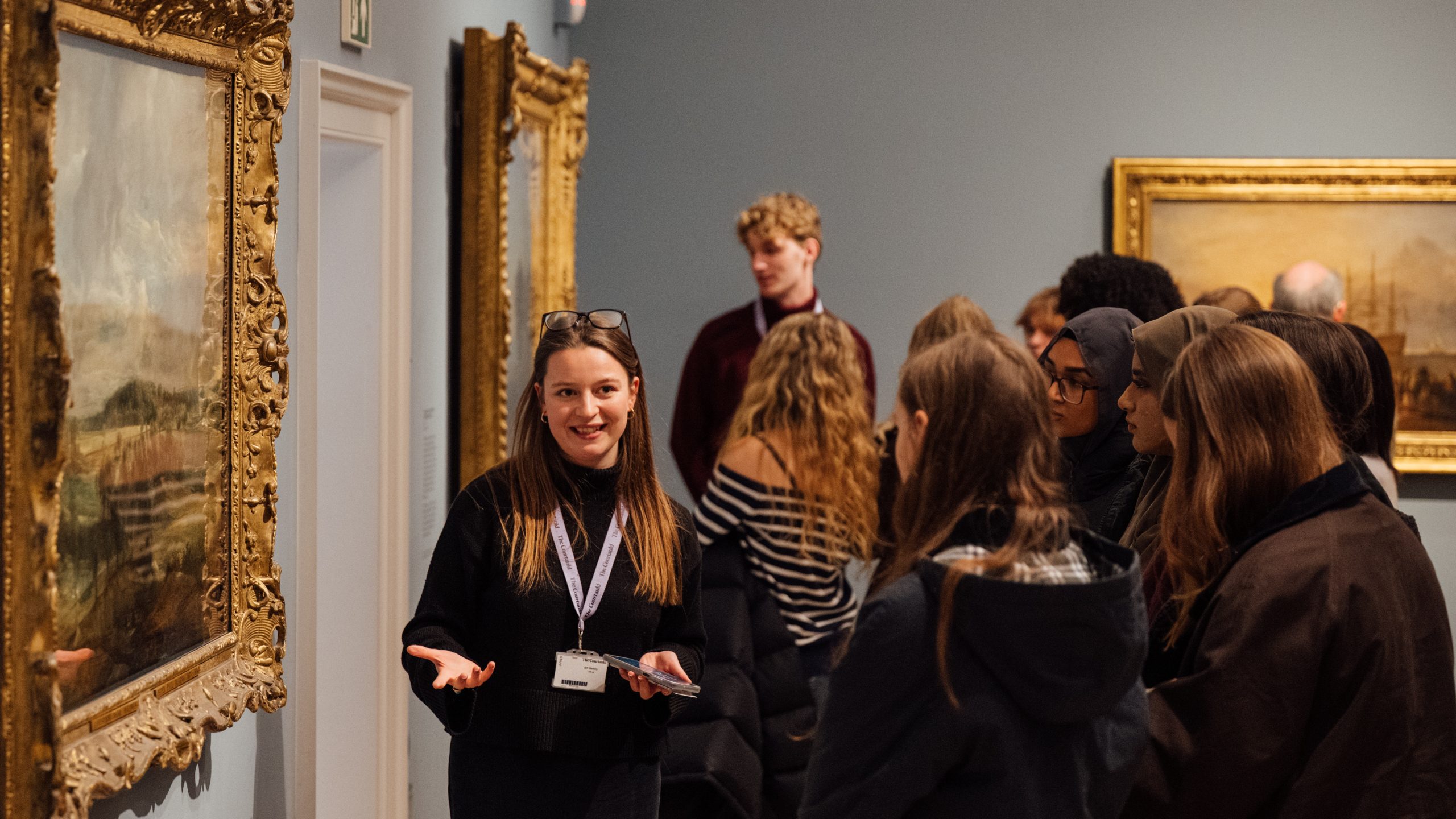 A teacher is stood by an artwork, speaking to a group of teenage school students.