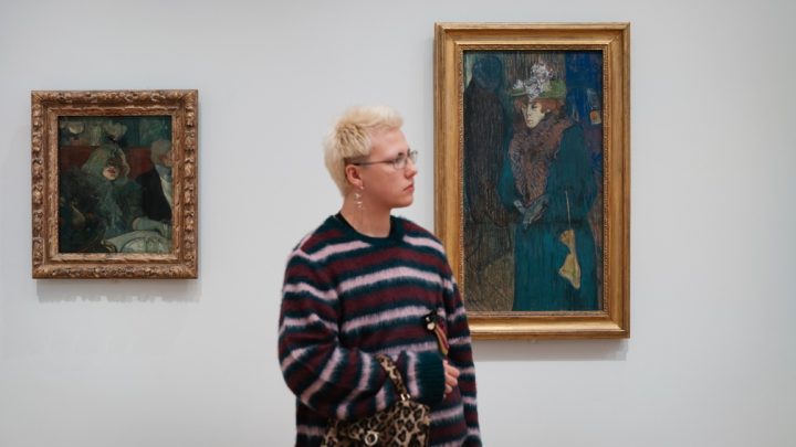 A young person with short bleach-blonde hair stands in the Courtauld Gallery in front of its white wall and between two works.