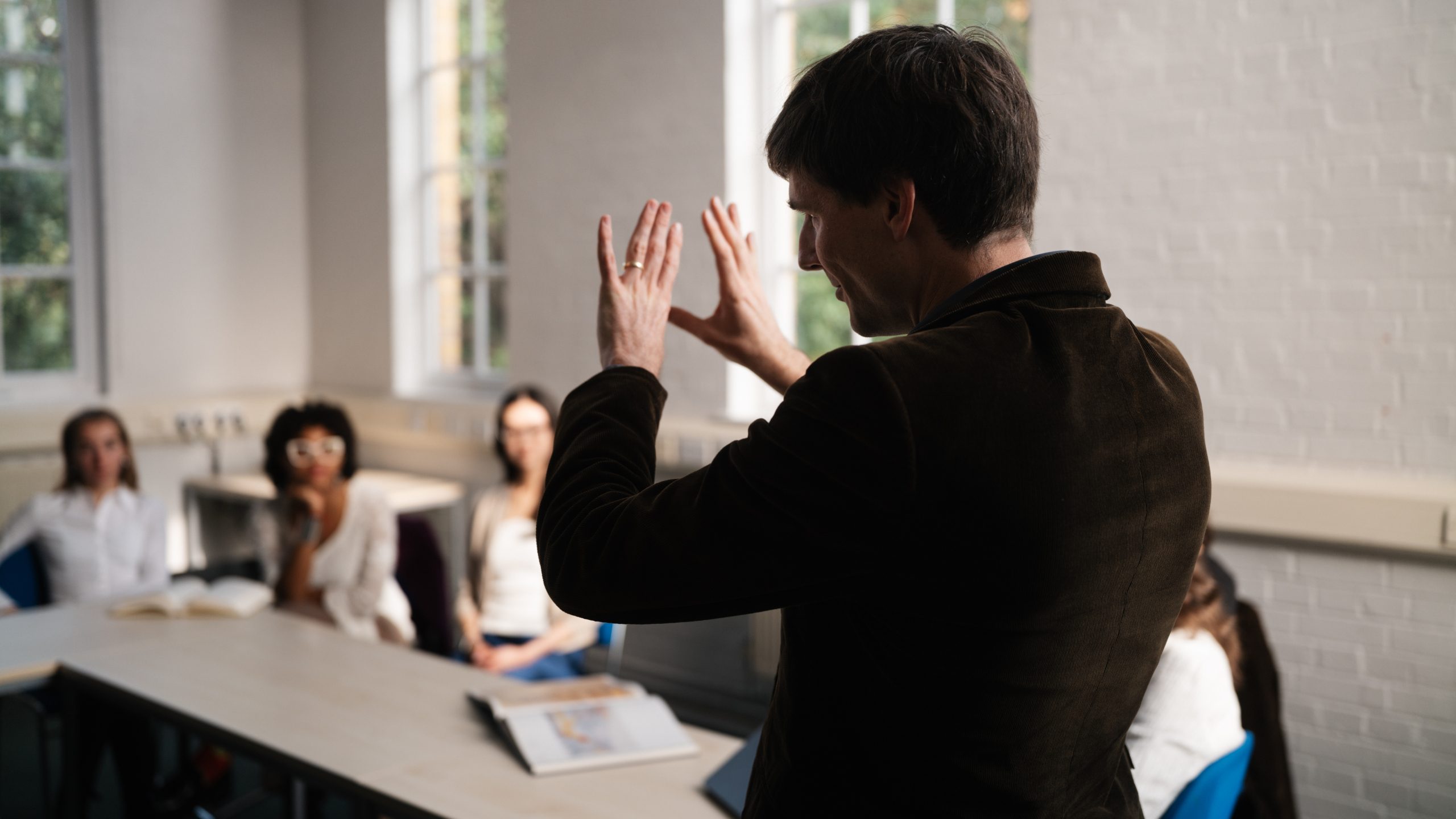 Tutor stands in front of class, his back to the camera. He is gesturing with his hands while students watch on.