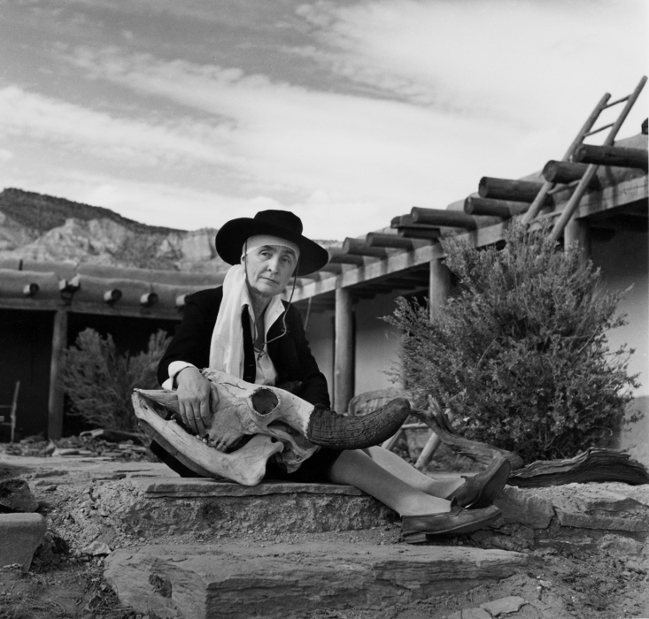 Black and white photograph of Georgia O'Keeffe at her home, with an open veranda patio visible in front of large mountains. She sits on a rock, holding a large deer's skull.