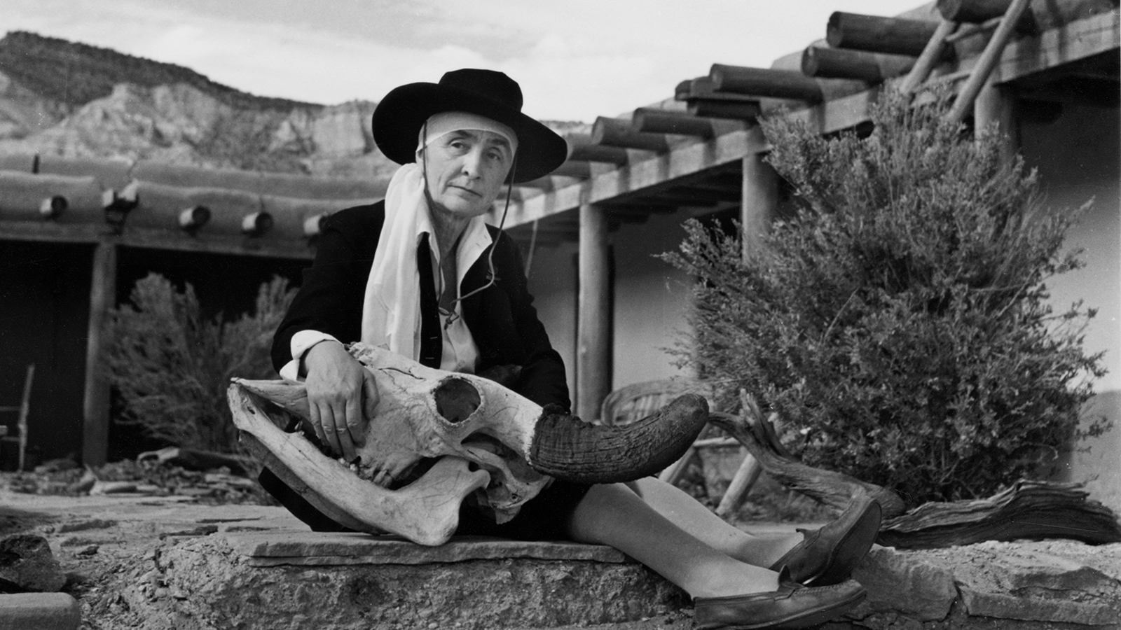 Black and white photograph of Georgia O'Keeffe at her home, with an open veranda patio visible in front of large mountains. She sits on a rock, holding a large deer's skull.