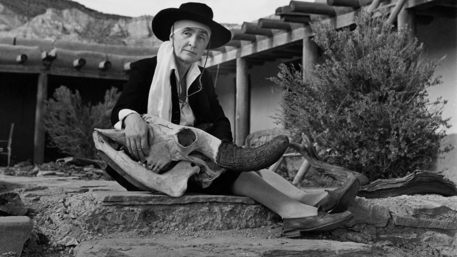 Black and white photograph of Georgia O'Keeffe at her home, with an open veranda patio visible in front of large mountains. She sits on a rock, holding a large deer's skull.