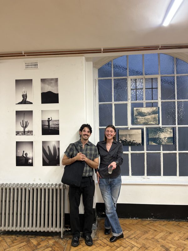 Two young people smile at the camera. They're stood between two arrangements of black and white photographs