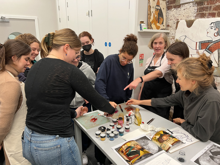 A group of students stand around a table which has pigments on it. Some of them are pointing and others are smiling. A woman with short hair wearing an apron is also pointing at the table.