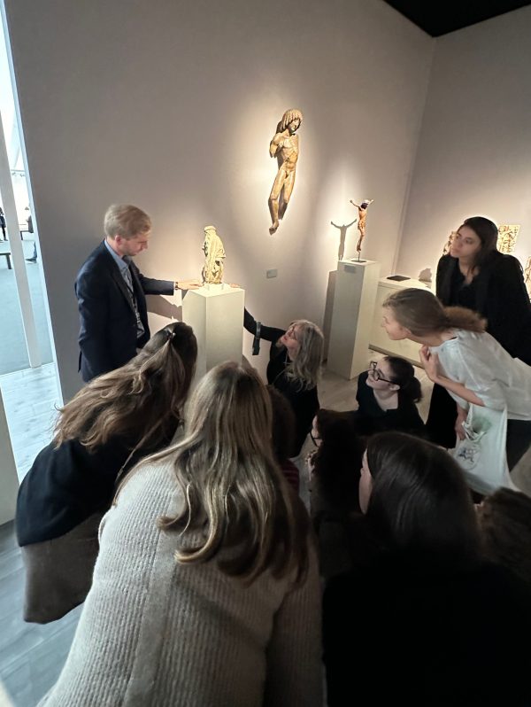 A group of students in a museum are gathered around a sculpture on a white podium, crouched and looking at it closely. A man stands beside the sculpture.