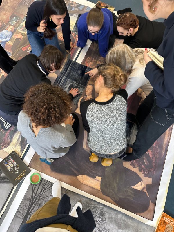 Students kneel on large prints of Renaissance art spread on the floor. They are pointing at and discussing details of the print
