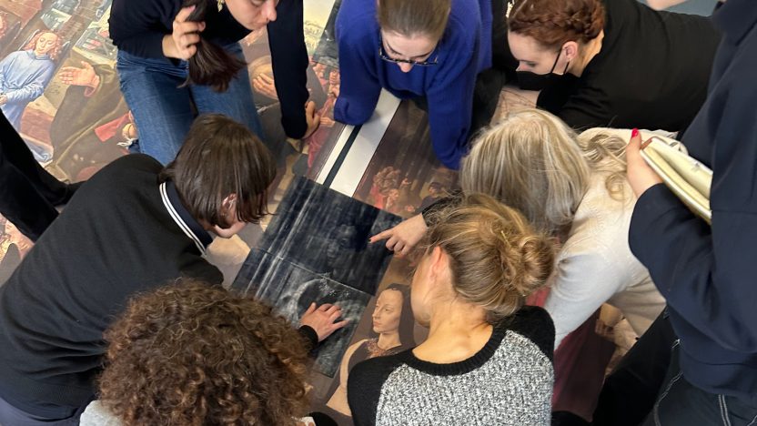 Students kneel on large prints of Renaissance art spread on the floor. They are pointing at and discussing details of the print