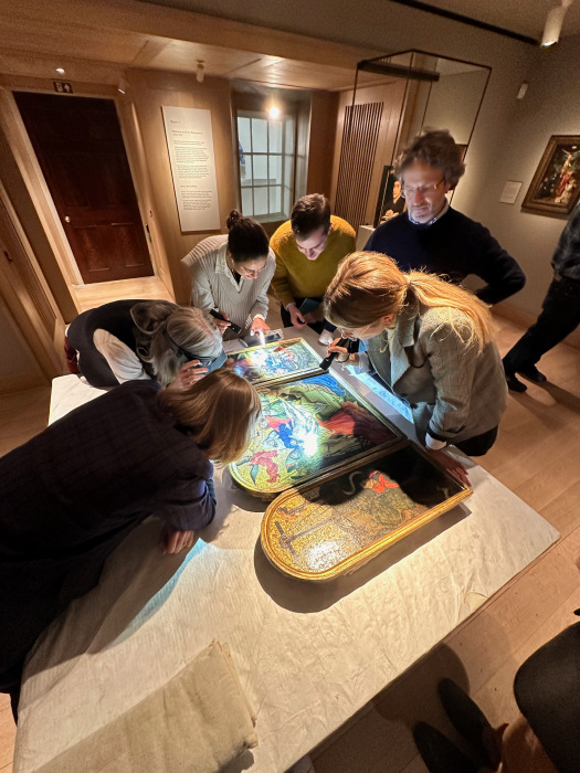 Six people are gathered around a triptych set on a table. Some of them have torches and are examining details. They are in a gallery