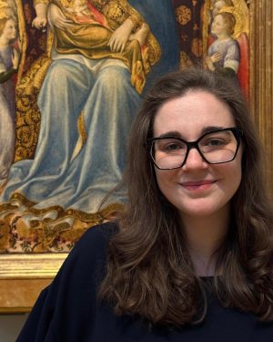 A woman with glasses smiles in front of the corner of an ornate medieval painting.