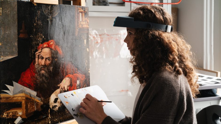 A woman with curly hair applies paint onto a canvas. She wears a headtorch.
