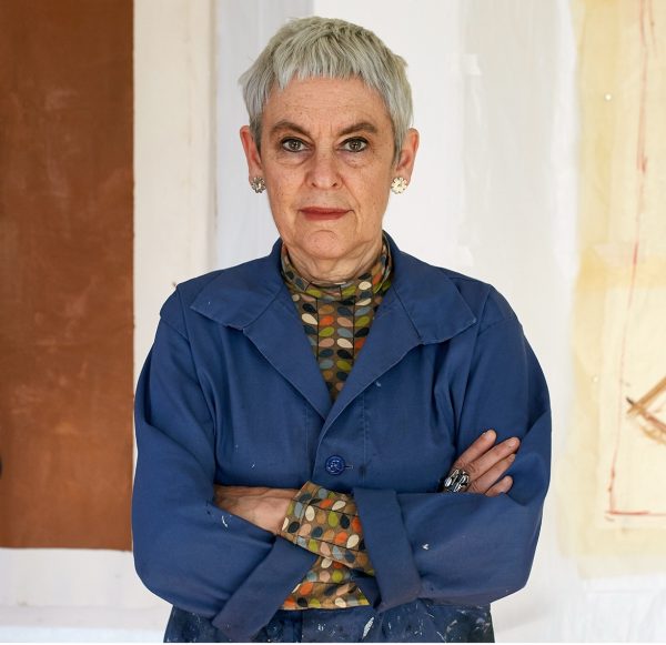 a woman with short grey hair stands arms folded for a portrait, she wears a blue shirt.