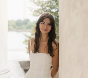 A woman in a white dress with long brown hair leans against a white column. Behind her is a lake and trees.