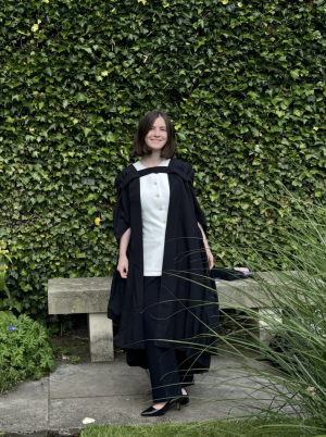 A woman with short brown hair in a degree gown stands in front of a wall of ivy. She smiles at the camera.