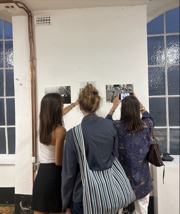 Three women gather around a display of three small photographs on the wall; one of them takes a photo on their phone, the other points at a photograph. The room is sparse and white with a visible copper pipe.