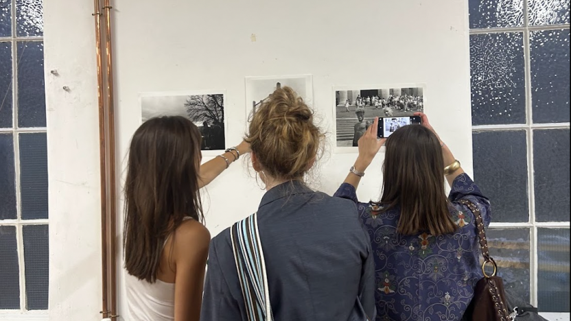 Three women gather around a display of three small photographs on the wall; one of them takes a photo on their phone, the other points at a photograph. The room is sparse and white with a visible copper pipe.