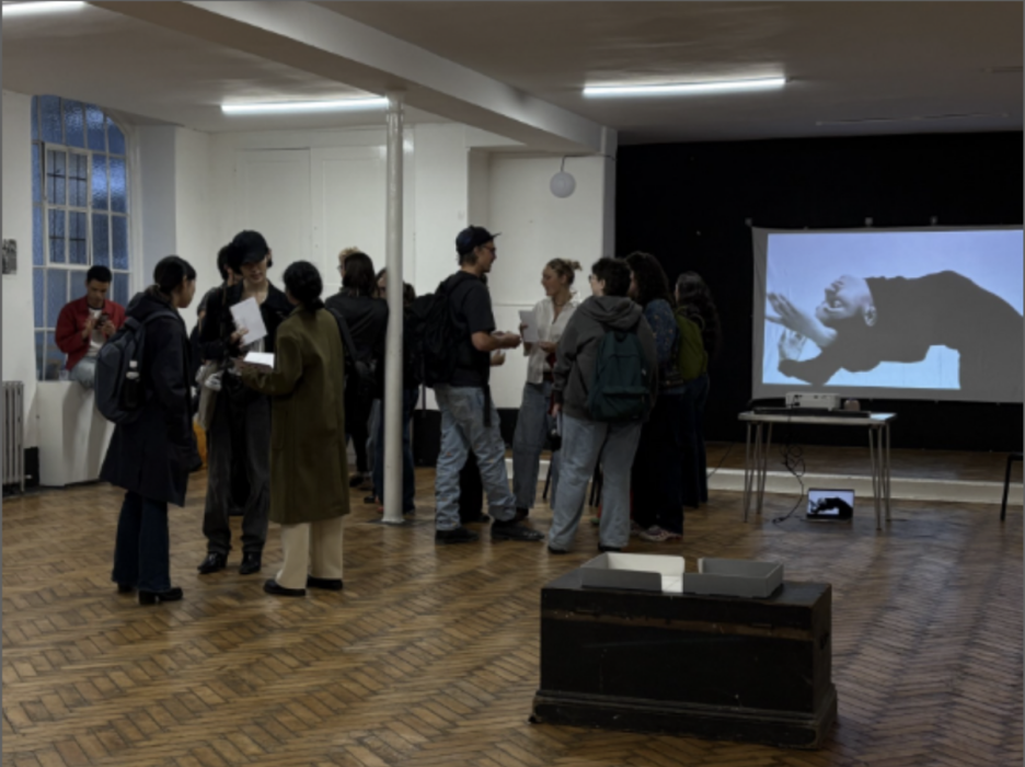 A group of people chatting holding catalogues in a big white room. A black and white film plays on a projector