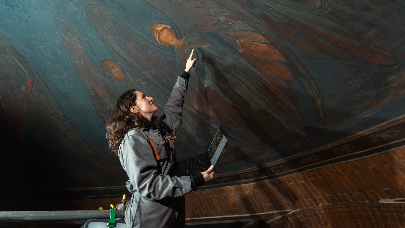 A woman in a jumpsuit points at paintings on a domed ceiling, painted with blue and orange winged women. She has a laptop in one hand and is tied to scaffolding just visible in the corner.