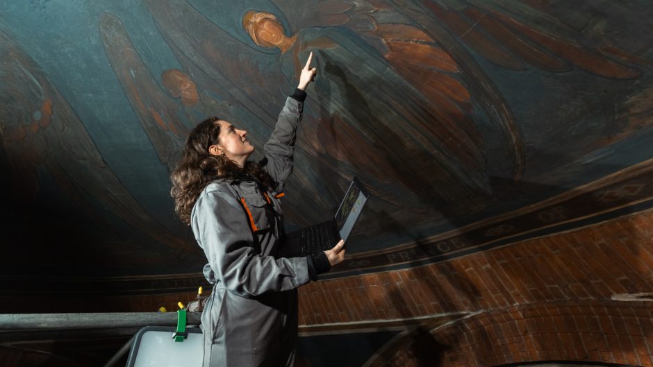 A woman in a jumpsuit points at paintings on a domed ceiling, painted with blue and orange winged women. She has a laptop in one hand and is tied to scaffolding just visible in the corner.