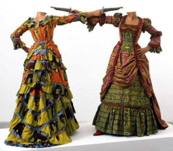 Two headless mannequins wear dresses like ornate 19th century European woman's dresses, constructed of vibrant, colorful fabrics reflect traditional African textiles. The two mannequin are raising rifles at each other.