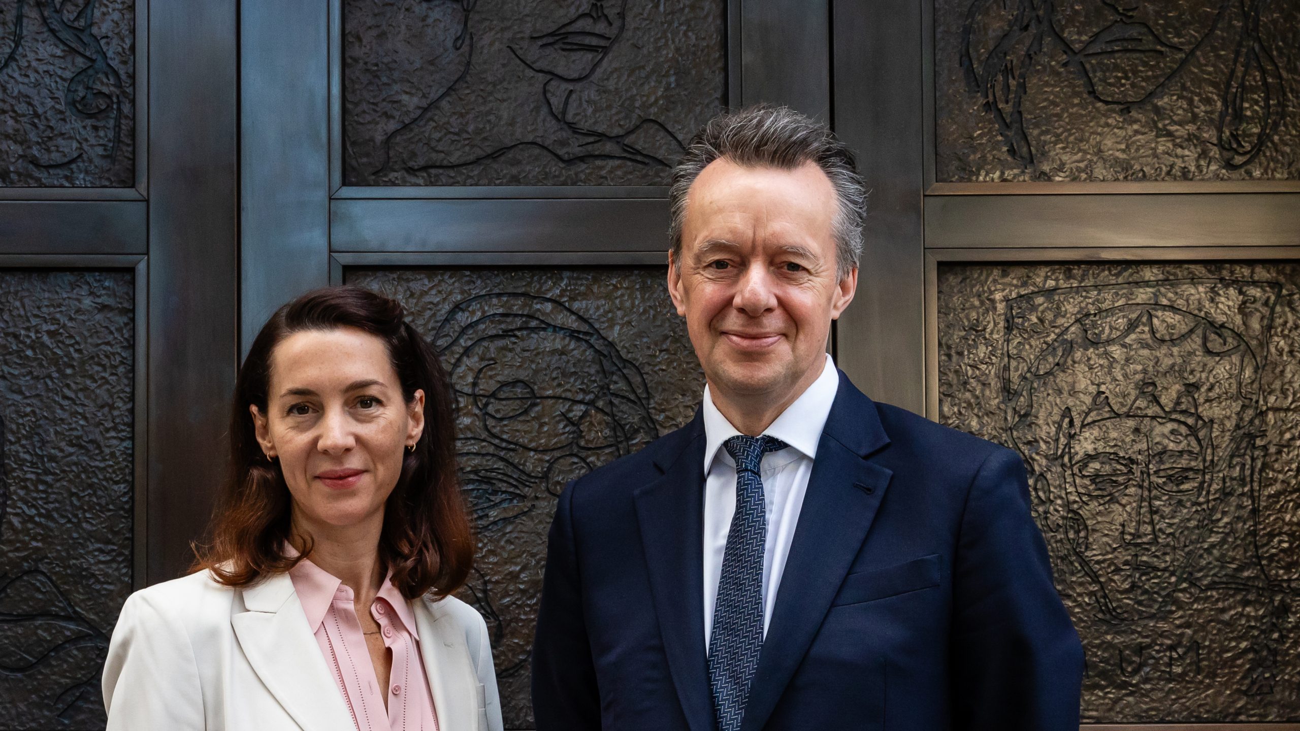 Two figures in suits stand in front of bronze doors