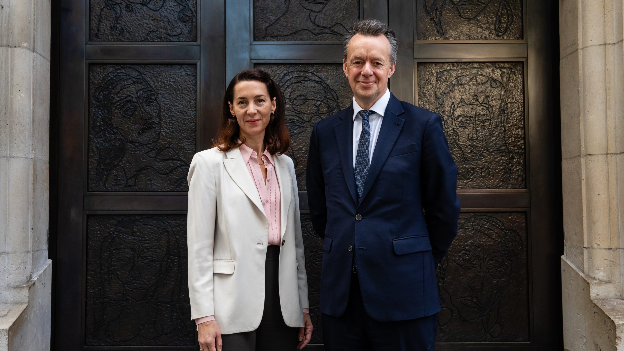 Two figures in suits stand in front of bronze doors